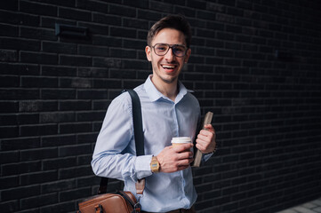 Smiling businessman holding coffee and book near brick wall