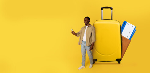 A traveler stands beside a bright yellow suitcase while using a phone with a travel app to plan...