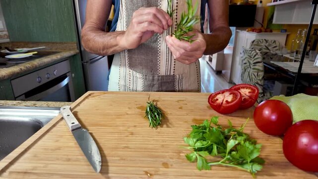 Experienced senior chef carefully arranging fresh rosemary sprigs while preparing vegetarian recipe, surrounded by vibrant tomatoes and parsley on wooden cutting board in home kitchen