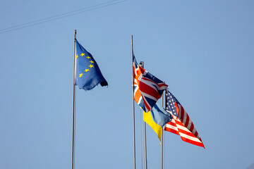 Flags of the European Union, United Kingdom, Ukraine, and the United States of America flying together on flagpoles against a clear blue sky.
