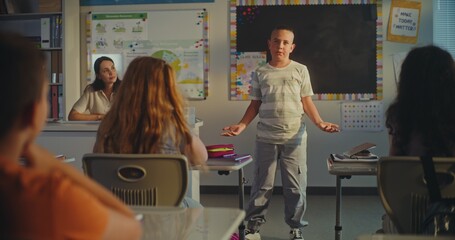 STEM Class on Renewable Energy: Elementary School Boy Presenting Homework on Ecology in Front of Classmates and Female Teacher. Diverse Children Sitting at Desks During Science Lesson in Classroom.