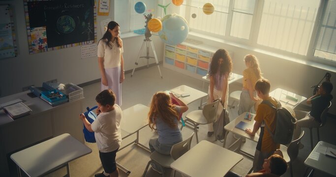 Female Teacher Greeting Boys and Girls at the Beginning of the School Day. Elementary School Students Entering Classroom, Sitting Down at Desks and Unpacking Their Backpacks. Slow Motion. High Angle.