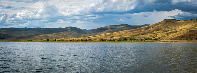Peaceful mountain lake near Gunnison, Colorado, with golden rolling hills and dramatic summer skies, reflecting the serene beauty and open landscapes of the American West.