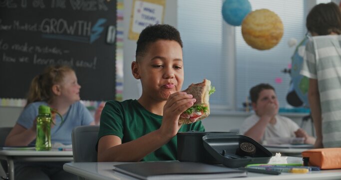 African American Primary School Boy Sitting at the Desk, Eating Sandwich from Lunch Box, Talking to Classmate During Break. Happy Diverse Kids Having Lunchtime Before Lesson in Multiethnic Classroom.