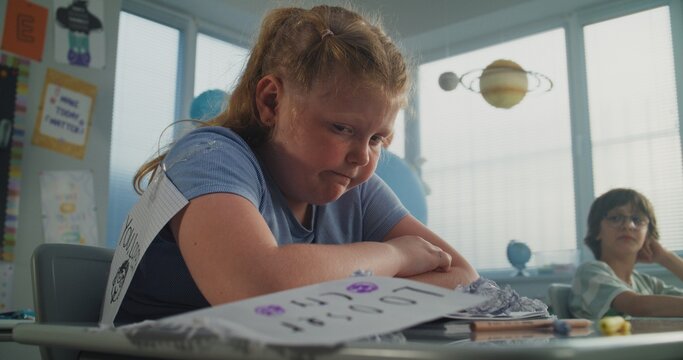 Upset Elementary School Student Sitting Alone at Desk and Crying While Classmates Bullying Her, Laughing and Throwing Papers. Girl Suffering from School Bullying, Peer Harassment and Emotional Abuse. - Powered by Adobe