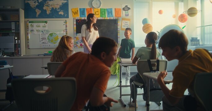 Female Teacher Introducing New Young Student in Front of Class Full of Elementary School Children. Group of Diverse Kids Sitting at the Desks, Clapping in Hands, Greeting New Classmate Before Lesson. - Powered by Adobe