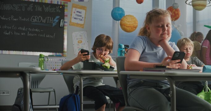 Primary School Children Eating Sandwiches, Using Smartphones and Talking with Each Other During Break. Happy Diverse Kids Sitting at Desks, Having Lunchtime Before Lesson in Modern Colorful Classroom.