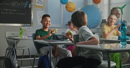 African American Elementary School Student Sitting at Desk, Eating Yummy Homemade Sandwich from Lunch Box, Talking to Classmate. Young Boys and Girls Having Lunchtime in Modern Classroom Before Lesson