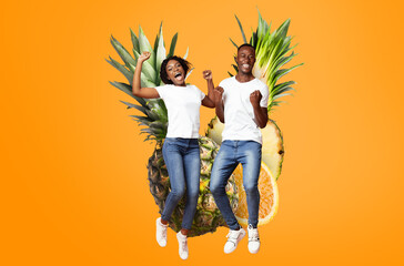 Two young African American adults jump in excitement against a bright orange background featuring pineapple and citrus images. Their expressions show pure happiness, embodying a fun summer vibe.