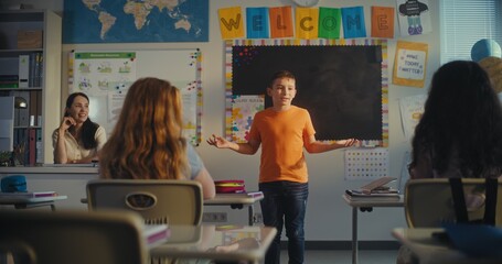 STEM Class on Renewable Energy: Female Teacher Calling Elementary School Student to the Board. Talented Boy Presenting Homework on Ecology in Front of Classroom Full of Diverse Children, Classmates.