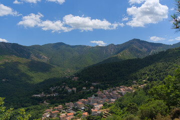 Obraz premium A charming village with red-tiled roofs nestles in a lush green mountain valley under a partly cloudy blue sky.