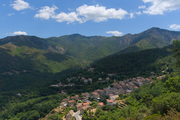 A charming village with red-tiled roofs nestles in a lush green mountain valley under a partly cloudy blue sky.