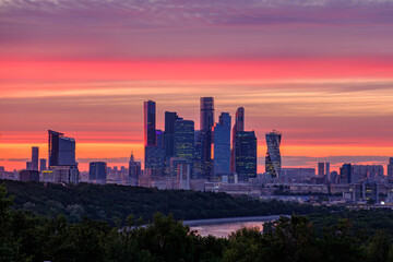 Panoramic view of Moscow City skyscrapers at sunset with vibrant orange and purple sky. Modern urban skyline with glass buildings, river, and park foreground. Beautiful evening in Moscow, Russia.