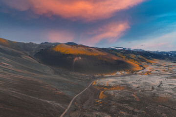 A volcanic crater with orange and yellow slopes, a winding dirt road, snow capped mountains, and...