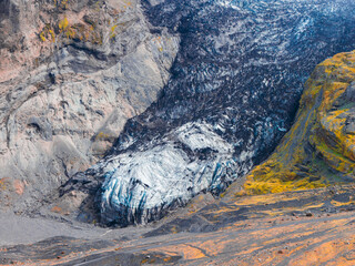 A glacier in Iceland with dark debris streaks lies against a rugged mountainside in Iceland, with yellow moss patches adding color to the stark terrain.