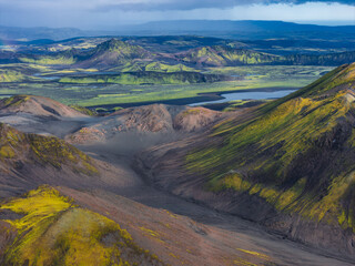 Aerial view of Iceland's rugged terrain with green moss, volcanic soil, craters, valleys, and a serene lake reflecting the surrounding landscape.