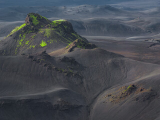 A volcanic terrain in Iceland features a moss covered rocky hill amid dark soil. Undulating hills and valleys are highlighted by soft, diffused light.