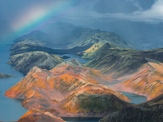 Aerial view of Iceland's rainbow mountains with multicolored terrain, a vivid rainbow in the sky, and serene lakes nestled among the hills.