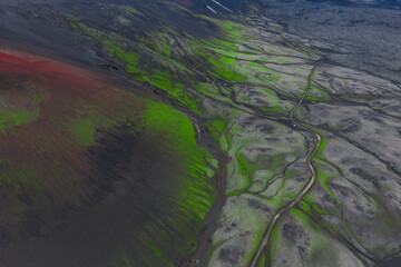 Aerial view of a red hued volcanic crater in Iceland, surrounded by green moss, dark soil, winding streams, and distant snow patches in the background.
