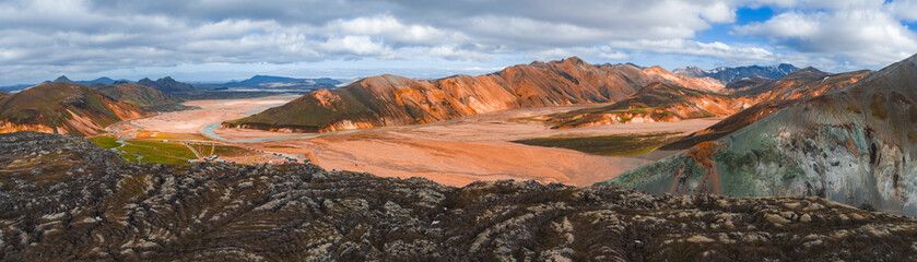 Vibrant orange, red, and green rhyolite mountains rise above a barren plain with mossy terrain, volcanic rocks, and a campsite in Landmannalaugar, Iceland.