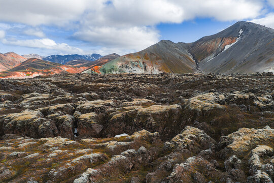 Volcanic lava fields in the foreground with colorful rhyolite mountains in the background, featuring orange, green, and gray hues under a partly cloudy sky.