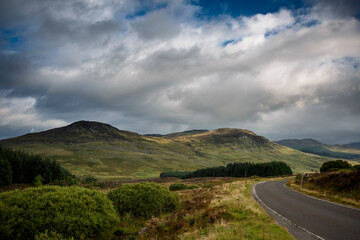Vast green hills and winding roads under dramatic clouds in a serene landscape of the Scottish Highlands during late afternoon light