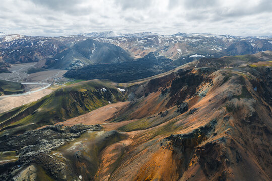 Aerial view of colorful rhyolite mountains in Iceland's Landmannalaugar region with snow patches, a winding road, and a dark lava field below. - Powered by Adobe