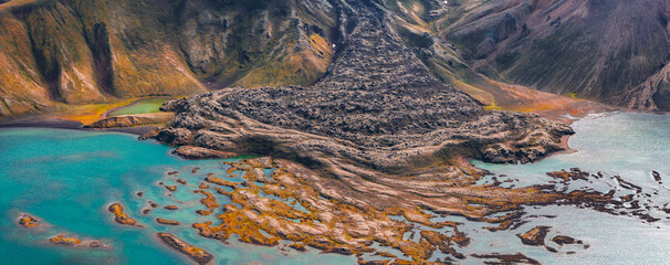 Aerial view of a volcanic terrain in Iceland with a lava flow leading into a turquoise lake, surrounded by multicolored hills and green moss.