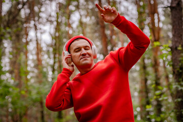 A man in a red sweater and a red beanie is dancing in a forest. He is wearing headphones and seems to be enjoying the music. The trees and greenery behind him create a beautiful and peaceful backdrop.