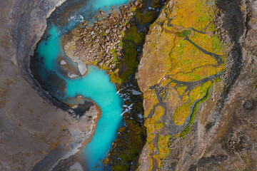 Turquoise river winds through rocky terrain and moss covered ground in Iceland. Aerial perspective highlights vivid colors and natural patterns.