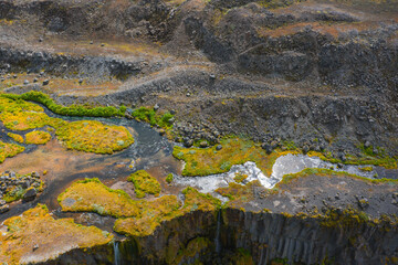 An aerial view of a rugged Icelandic landscape with a narrow waterfall cascading over a cliff, bordered by basalt columns, moss, and golden vegetation.