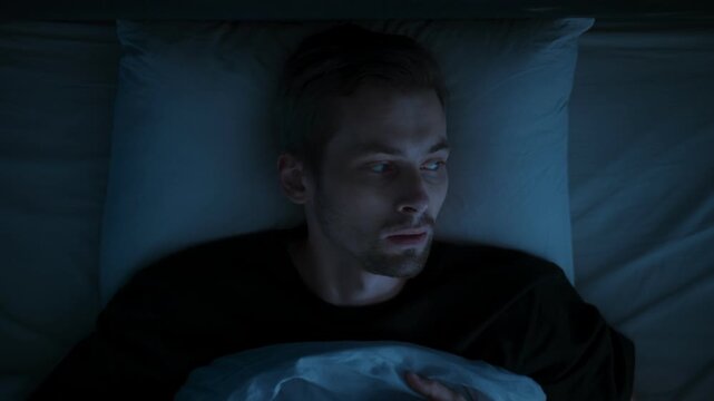 Close-up, in a dark bedroom at night, a man wakes up because of sleep paralysis or nightmare, drained by insomnia and a sleep disorder, overwhelmed by anxiety, fatigue.
