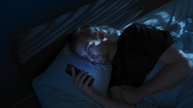 A sleepless young man lies awake in his bedroom at night, using his smartphone scrolling social media. Digital dependence and insomnia concept. Space for text