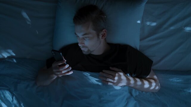 Top view, young man laying in bed at night, holding smartphone and doom scrolling through social media, then tossing the phone aside in exhaustion and frustration. Smartphone addiction and insomnia