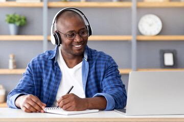 E-education concept. Happy black mature man using laptop and taking notes, listening online course...