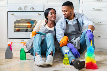 Tired young black couple sitting on floor among cleaning supplies after finishing home cleanup,...