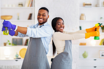 Portrait of young black couple using detergent spray bottles as guns, posing during cleanup at...