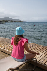 A girl sits cross-legged on a lounger, facing the sea in meditation