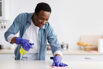 Smiling black guy cleaning kitchen furniture, using spray bottle with disinfestor and dust cloth,...
