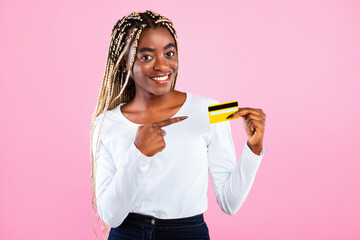 Pretty young African American woman pointing at yellow bank credit card, pink studio background. Contactless payment, easy banking