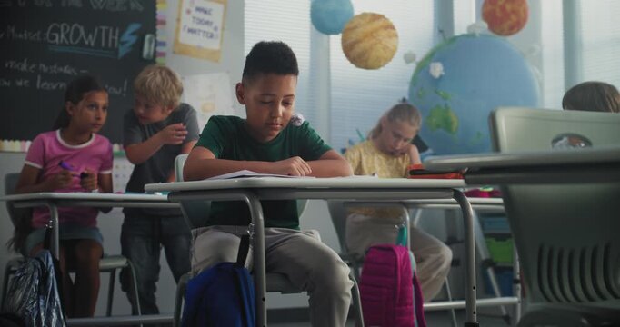 Stressed African American Primary School Boy Sitting Alone at Desk While Aggressive Classmates Abusing Him, Throwing Papers and Laughing. School Bullying, Racial Harassment, Toxic School Environment.