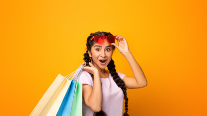 Happy Customer. Portrait of excited emotional young indian woman wearing and touching sunglasses holding colorful shopping bags with gifts, amazed lady looking at camera, yellow studio background