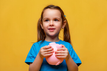 Happy little girl in blue t-shirt holding pink piggy bank money box and looking on yellow background. Financial concept of children pocket money. Save money for education and family expenses