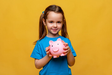 Happy little girl in blue t-shirt holding pink piggy bank money box and looking on yellow background. Financial concept of children pocket money. Save money for education and family expenses