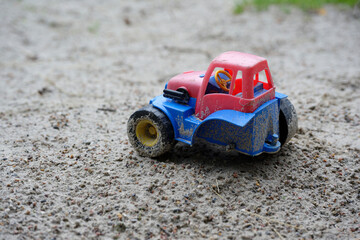 In the sand of a sandbox is a blue toy, a tractor with a large rear wheel missing