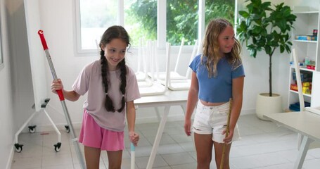 Two young diverse girls are tidying up a classroom or playroom. They have placed chairs upside down on the tables and are sweeping and cleaning the floor