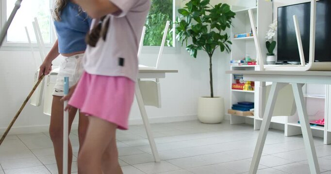 Two young diverse girls are tidying up a classroom or playroom. They have placed chairs upside down on the tables and are sweeping and cleaning the floor