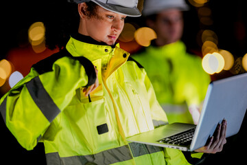 A focused female engineer in safety gear works on a laptop at night. Concentrated technician analyzing data or programming on-site with blurred industrial lights in the background.