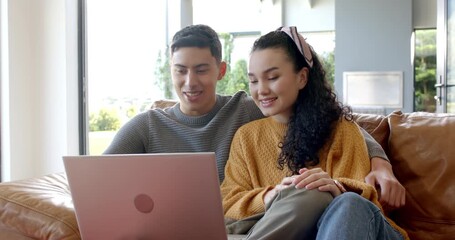 Diverse couple sitting on leather couch by window opening silver laptop and browsing pages together - Powered by Adobe
