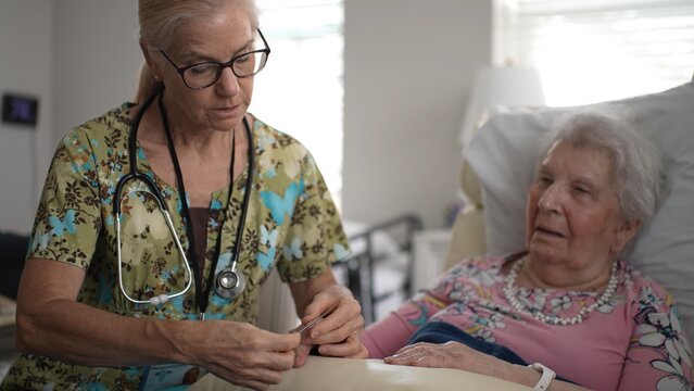 A nurse carefully files the nails of an elderly woman sitting in bed. The atmosphere is calm as they share a caring moment.